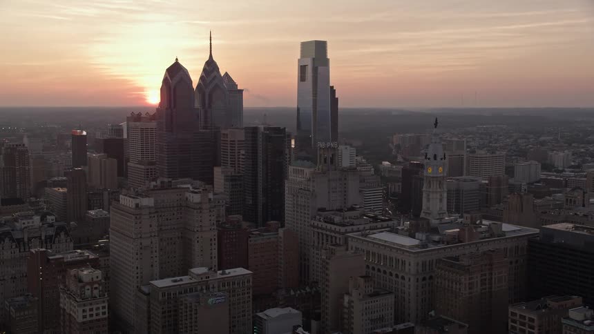 Aerial footage gliding over Philadelphias downtown skyscrapers as the sun sets behind them capturing the hazy metropolis at dusk