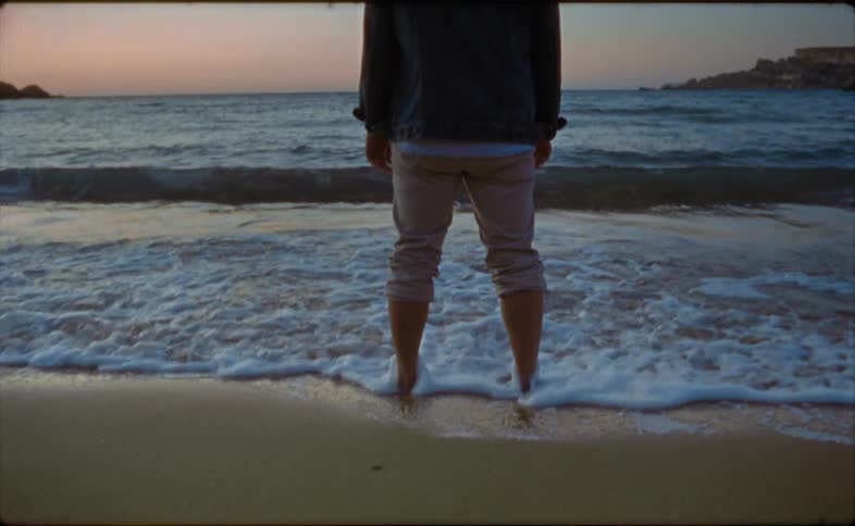 A man is standing barefoot on the beach with his pants rolled up as the waves roll over his feet