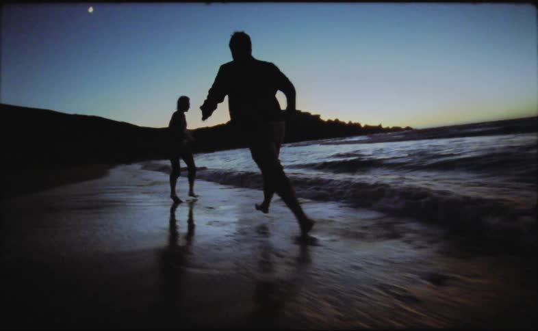 Man and woman running on the beach near the ocean at dusk are silhouetted against the sky