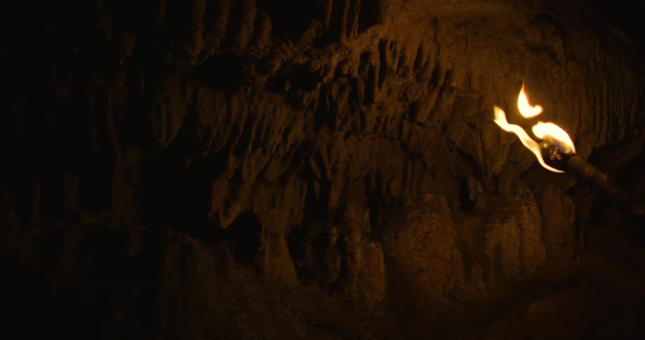 The flame of a torch burns brightly illuminating the walls inside a cave as a man makes his way though the darkness