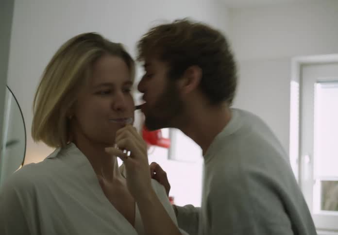 A man and a woman are standing in the bathroom together each holding a toothbrush in their mouth as they brush their teeth as he kisses her on the cheek