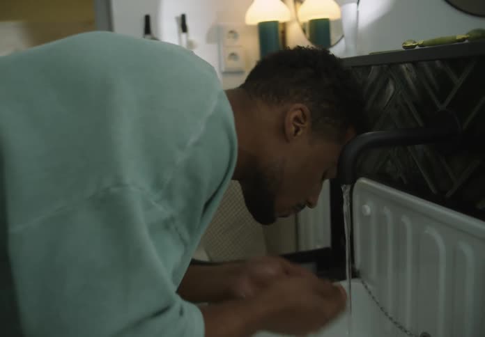 A man is standing over the bathroom sink as he turns on the faucet and splashes water on his face with his hands