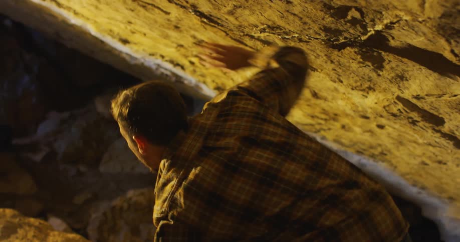 Man puts hand on low cave ceiling as he moves up slope
