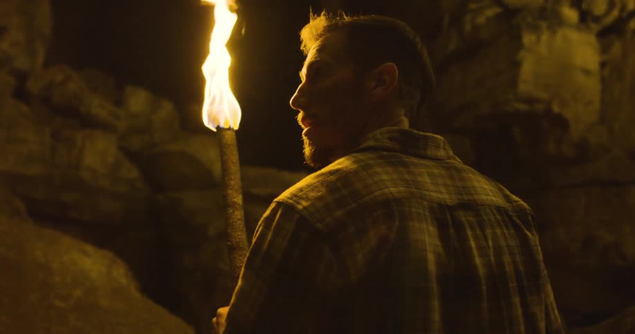 A man holding a fire torch looking nervously at a rock face