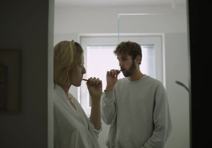 A man and a woman are standing in the bathroom together each holding a toothbrush in their mouth as they brush their teeth