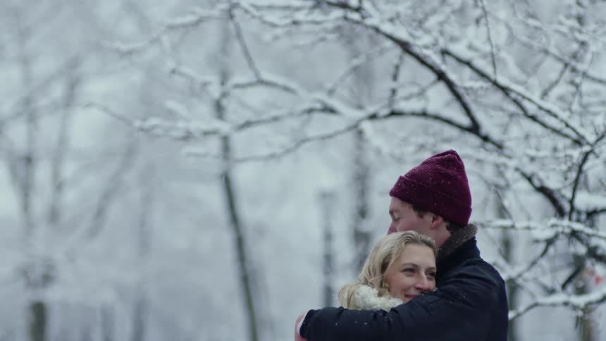 A man wearing a winter coat and a knit cap has his arms around his girlfriend as they stand outside hugging near snow covered trees as it snows on a winter day
