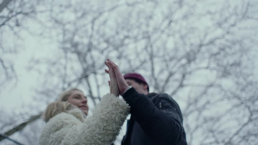 Couple touch hands while standing outdoors under leafless trees during snowfall