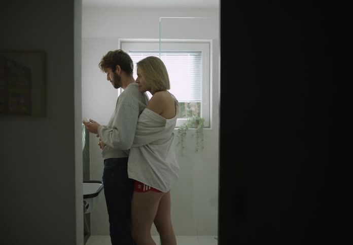 A woman walking up to a man standing in the bathroom and hugging him from behind