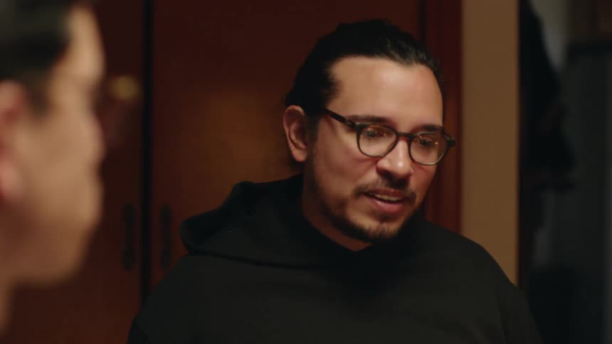 Man with glasses beard and black hoodie sits at dining table talking to friends in room