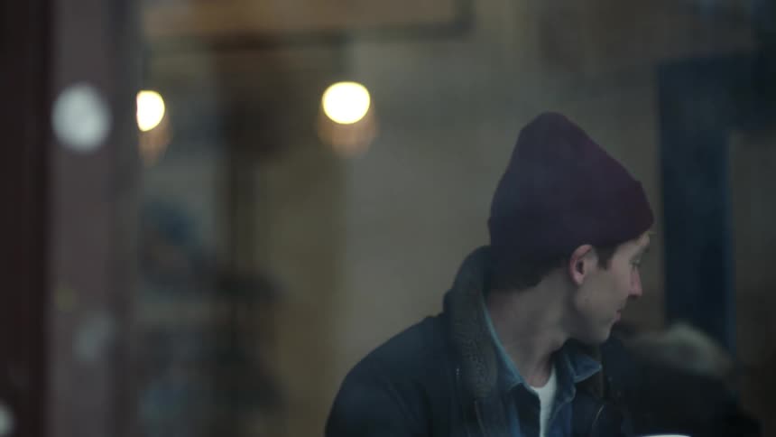 Young man wearing a beanie sits on a restaurant table near a window looking at people