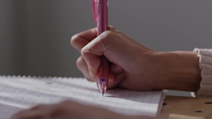 A woman is sitting at a table holding a pink pen as she writes in a spiral notebook