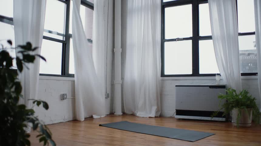 A room with bright windows white curtains blowing in the wind and a yoga mat on the wood floor