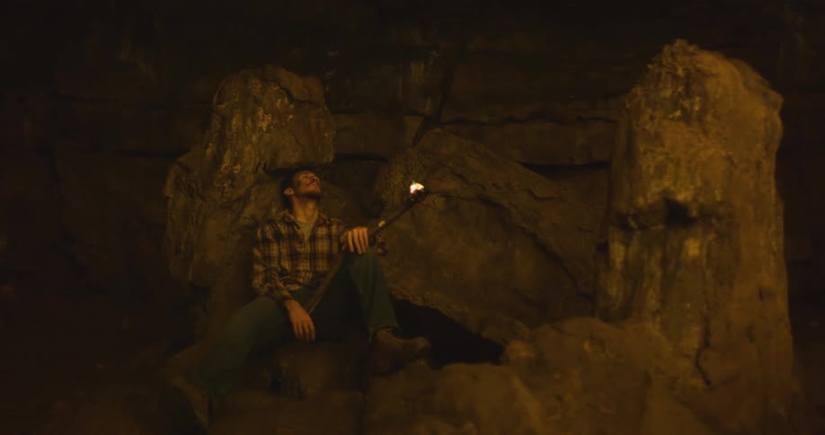 A man is sitting on rocks inside a dark cave looking up as he holds his torch with a dim flame