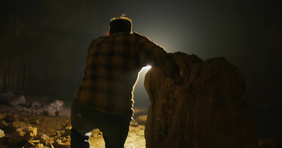 A man is moving past rocks through a cave as he holds his hand up to block the bright light coming in from outside