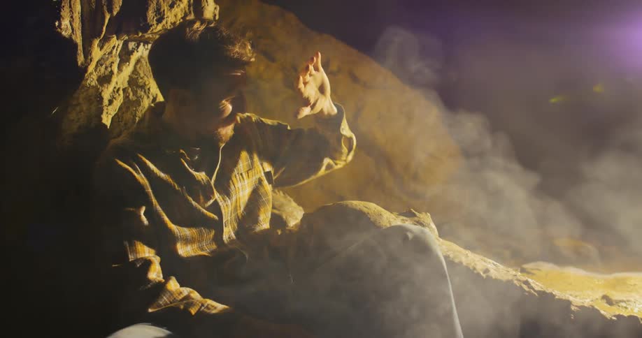 Man with beard green shirt and pant sits near large rock and light flashes in his eyes all of a sudden