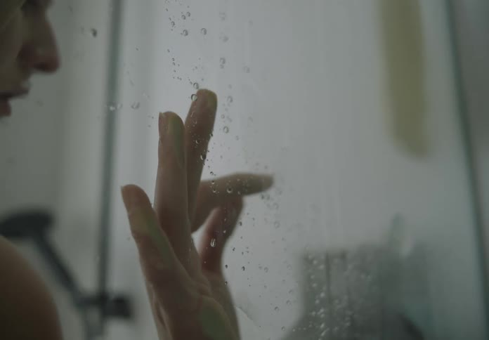 Person standing in shower stall holds hand against damp glass