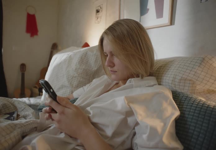 A young woman is laying down in her bed and is on her phone