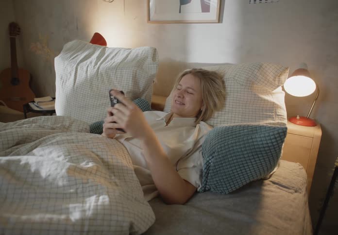 Blonde woman with white shirt lies in bed using smartphone and laughing in bedroom