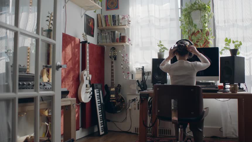Man with bun and white shirt sitting at desk wears headphones and uses computer in room