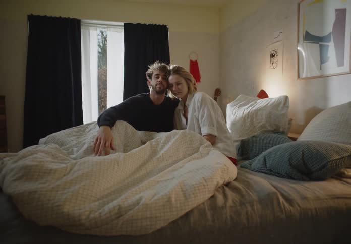A man and a woman are sitting up in bed together as his hand is on the blanket
