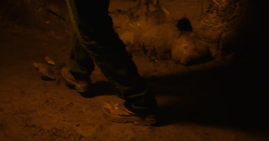 A man is walking slowly at night while carrying a torch along a dirt path next to a stone wall