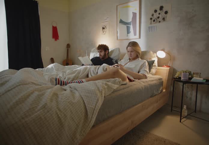 A man and woman sitting in bed the woman on her phone and the man reading a book