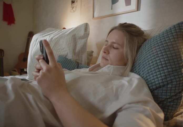 A woman is lying in bed holding her cell phone with both hands and she smiles