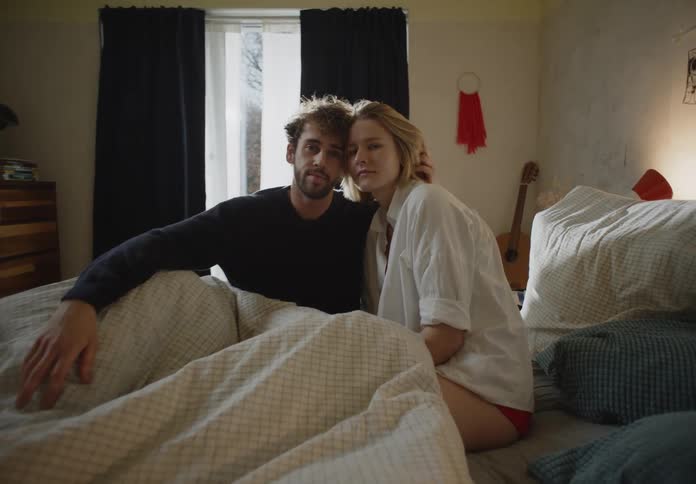 Man with beard and black sweater and blonde woman in white shirt and pink panties sit on bed posing for the camera in bedroom