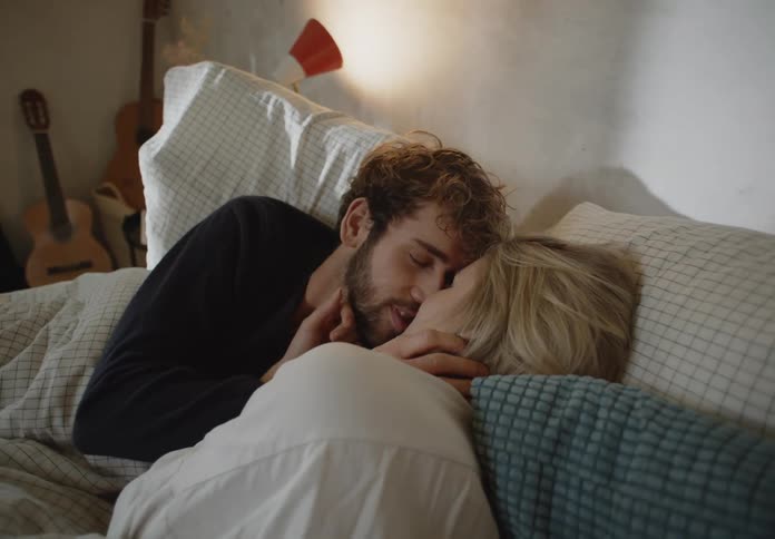 Man with beard black sweater and woman in white shirt lie in bed making out in bedroom