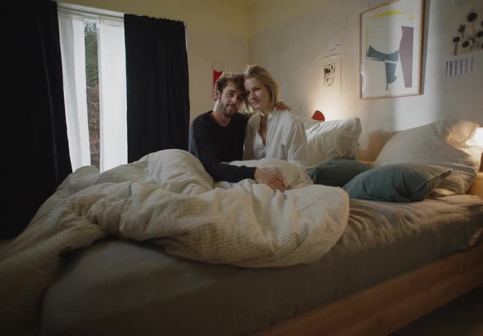 Man with beard and black sweater and blonde woman in white shirt and pink panties sit on bed posing for the camera in bedroom