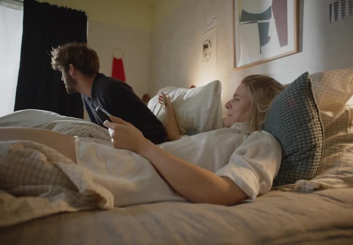 A woman is lying in bed texting on her phone as her husband reaches over and kisses her and they hug