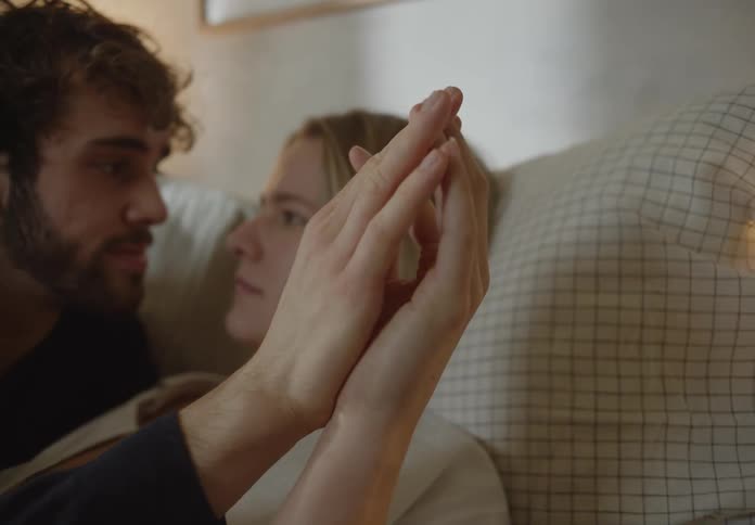 Man with beard and black sweater and blonde woman in white shirt lovingly looks into each others eyes holding hands on bed in bedroom