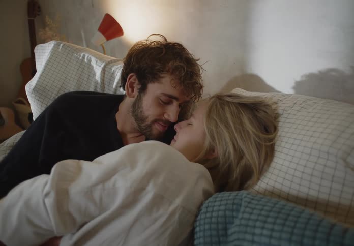 Man with beard and black sweater and blonde woman in white shirt make out in bed smiling in bedroom