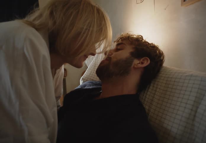 Man with beard and black sweater and blonde woman in white shirt make out in bed in bedroom