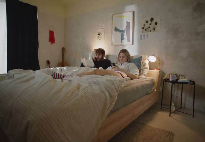A man and woman both recline in bed while the man reads and the woman looks at her phone