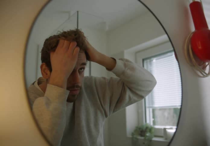 A man is standing in front of the bathroom mirror moving his hair with his hand as he looks at his face
