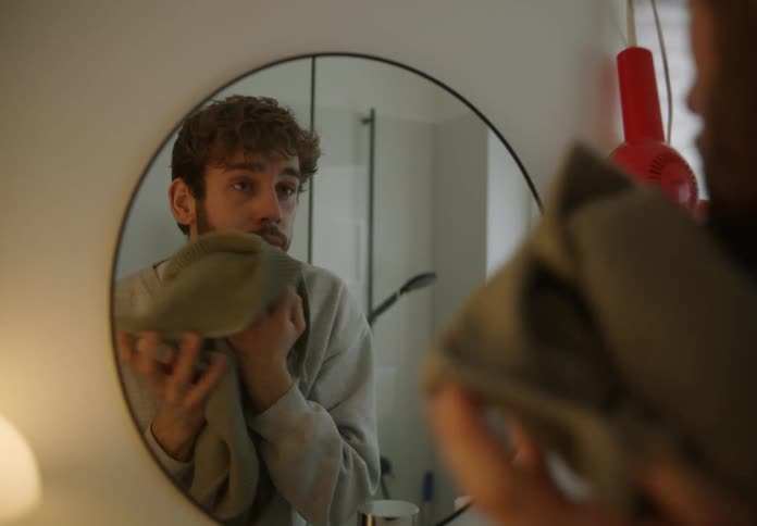 A young white man is wiping his face in the mirror