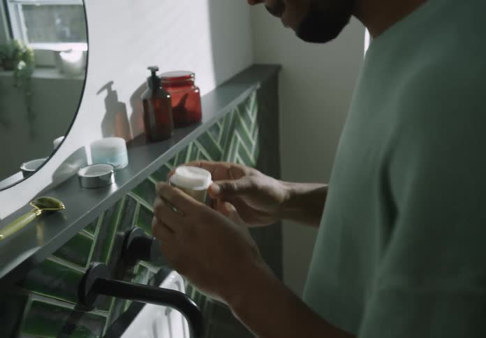 A man opens a jar of cream and applies it to his face while looking in a bathroom mirror