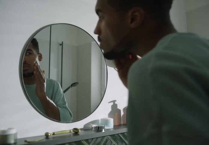 Black man applies lotion to his face while looking in a bathroom mirror