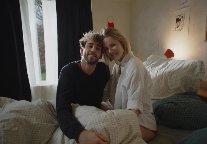 A man and woman smile into the camera while sitting with their heads pressed together in bed