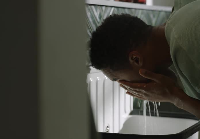 A man is standing over the bathroom sink as he splashes water on his face with his hands