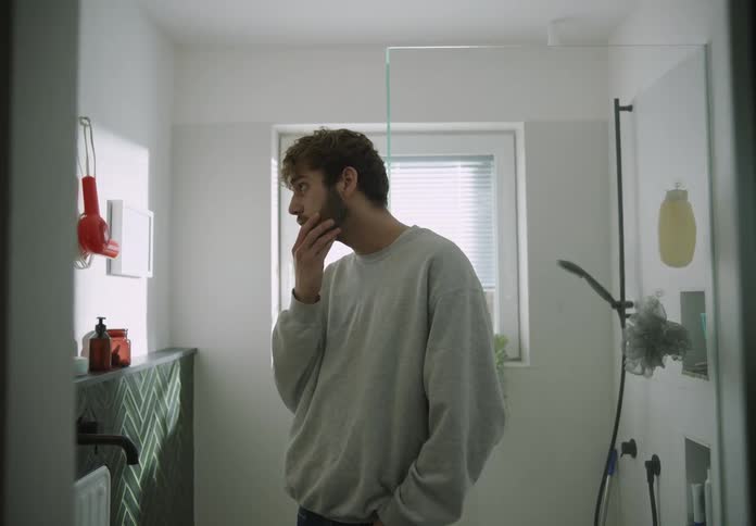 A man is standing in the bathroom as he turns his head to look at himself in the mirror and rubs his hand on his beard and turns with a smile