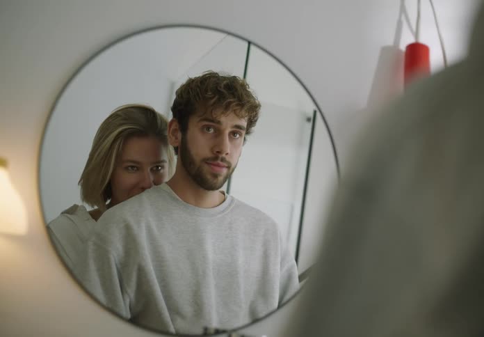 A man and woman stand while looking in a bathroom mirror