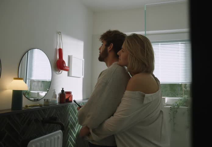 Man with beard and gray sweater brushes his teeth and blonde girlfriend comes to cuddle him in the bathroom