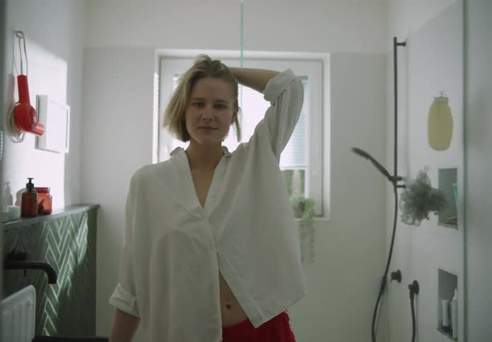 A woman wearing a white shirt is standing in the bathroom with her hand on her head as she moves her blond hair