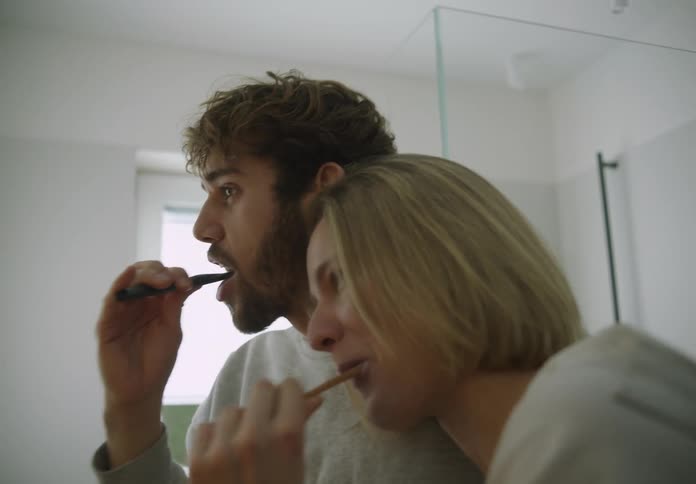 A man and a woman are standing in the bathroom together each holding a toothbrush in their mouth as they brush their teeth
