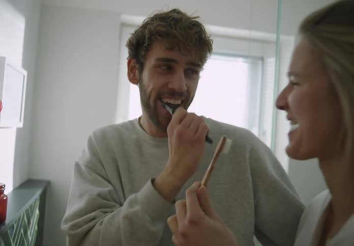 A young couple playfully brush their teeth in front of a bathroom mirror in the daytime