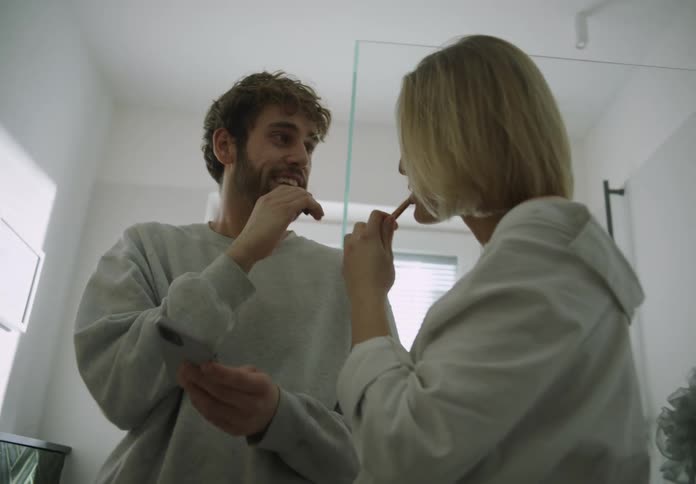 A man and woman stand in the bathroom brushing their teeth talking and looking at their phone