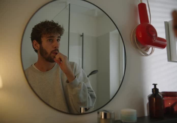 A man is standing in front of the bathroom mirror as he is holding a toothbrush and brushes his teeth