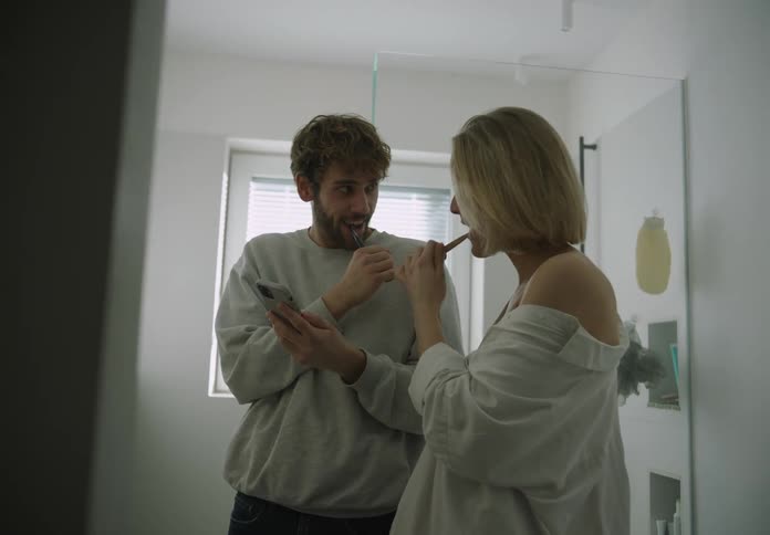 A man and woman talk and look at their phone while brushing their teeth in a bathroom